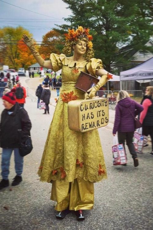 Woman all in gold and on stilts holds a suitcase which has the words "Curiosity has its rewards" printed on it.